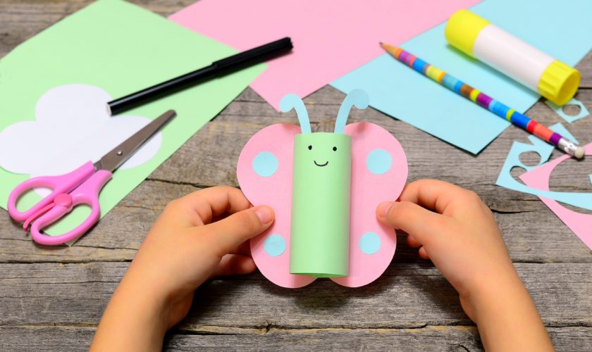 Small child holding paper butterfly in his hands. Child shows paper crafts. Funny butterfly made of colored paper. Office supplies on a wooden table. Children creativity concept