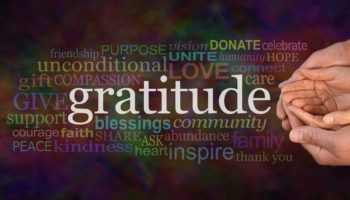 Gratitude Attitude Website Campaign Banner –  Male hands  cradling female cupped hands on a wide warm dark multicolored background with a GRATITUDE word cloud