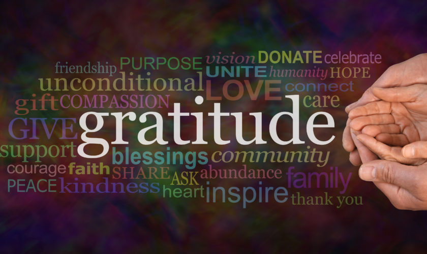 Gratitude Attitude Website Campaign Banner –  Male hands  cradling female cupped hands on a wide warm dark multicolored background with a GRATITUDE word cloud